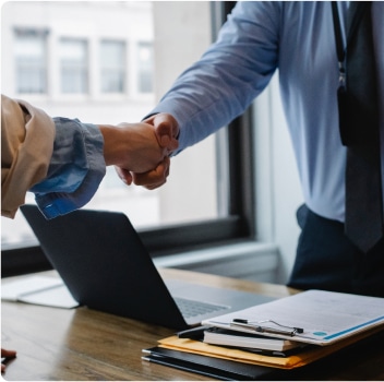 Two professionals shaking hands over a desk in an office setting.