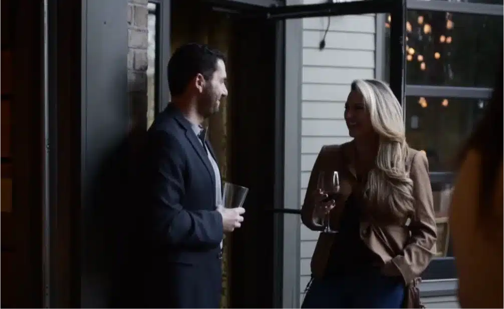 Two people chatting and drinking wine indoors near a window.