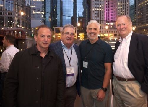Four men posing together at a nighttime outdoor event with city lights in the background.
