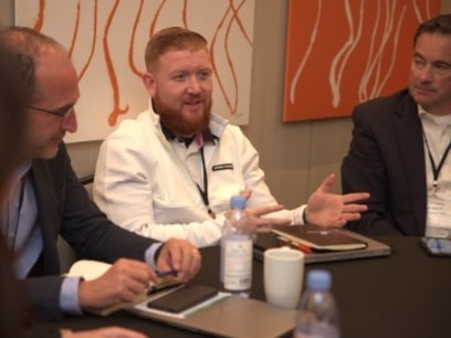 A man in a white jacket speaks at a meeting with others.